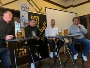 Die Gäste des Krombacher FanTalks – Thorsten „Fichte“ Friedrich, Andreas Klement, Sebastian Schneider und Jan Wilhelm – sitzen nebeneinander auf Barhockern, vor ihnen Biergläser auf Stehtischen. Jan Wilhelm spricht ins Mikrofon. Im Hintergrund ist ein VfL Eintracht Hagen Banner zu sehen.