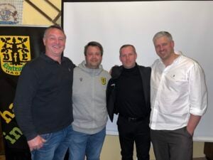 Gruppenfoto beim Krombacher FanTalk des VfL Eintracht Hagen am 26. März 2025: Thorsten „Fichte“ Friedrich, Jan Wilhelm, Moderator Andreas Klement und Sebastian Schneider stehen nebeneinander vor einer Leinwand und dem Vereinsbanner, lächeln in die Kamera.