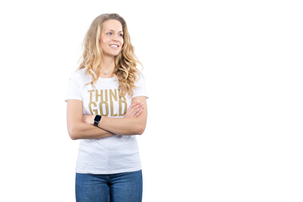 Christina Honsel steht mit verschränkten Armen vor einer weißen Wand. Sie trägt ein weißes T-Shirt mit der goldenen Aufschrift „THINK GOLD“ und blickt selbstbewusst in die Kamera.