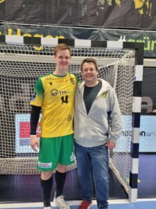 Zwei Personen stehen nach einem Handballspiel lächelnd vor dem Tor; einer trägt ein gelb-grünes Trikot mit der Nummer 14. Ein persönlicher Moment voller Wertschätzung und Leadership.